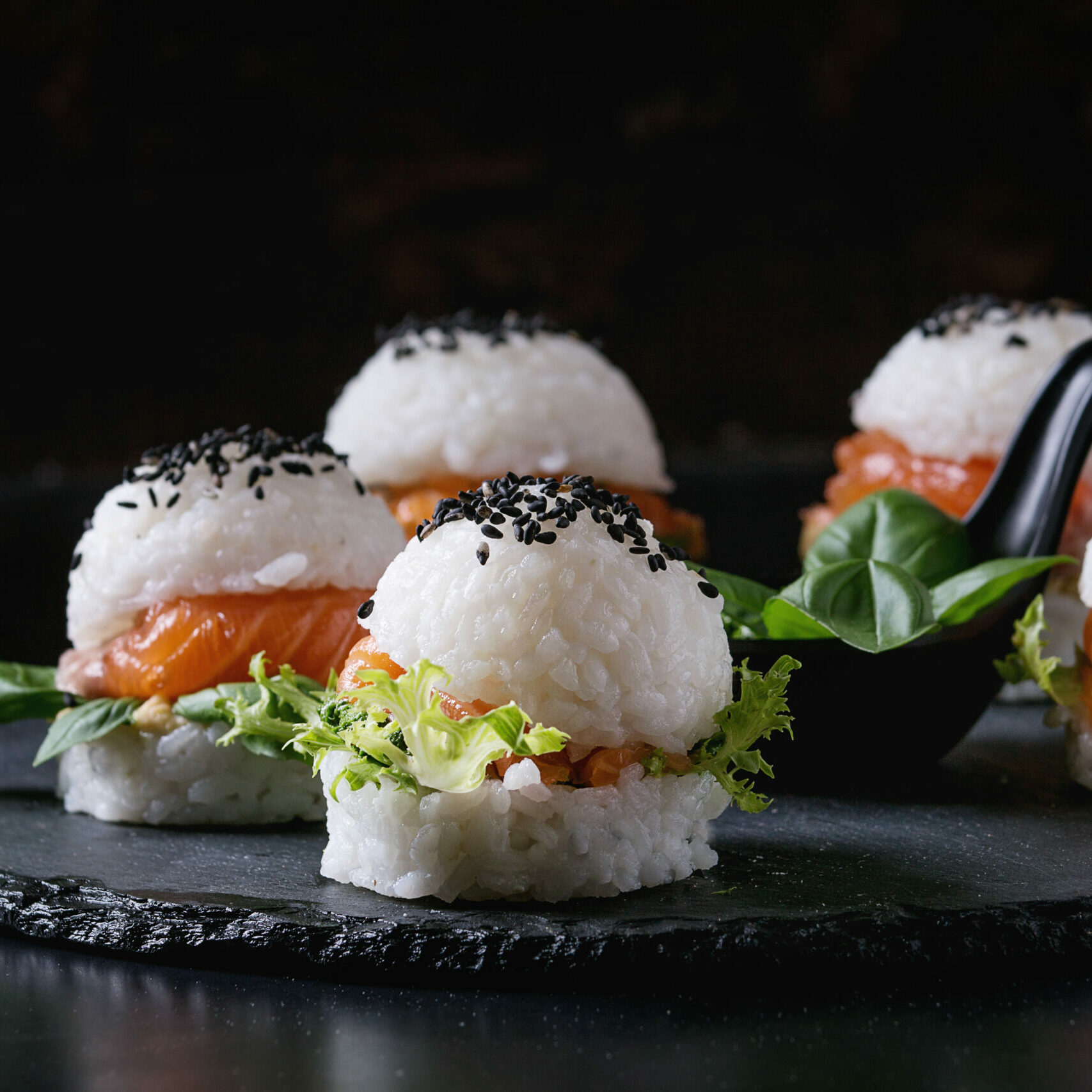 Mini rice sushi burgers with smoked salmon, green salad and sauces, black sesame served on slate stone board over black background. Modern healthy food